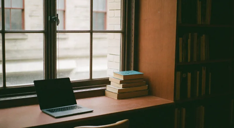 A wooden reading desk in a library alcove with an open laptop and a stack of books in diffused daylight