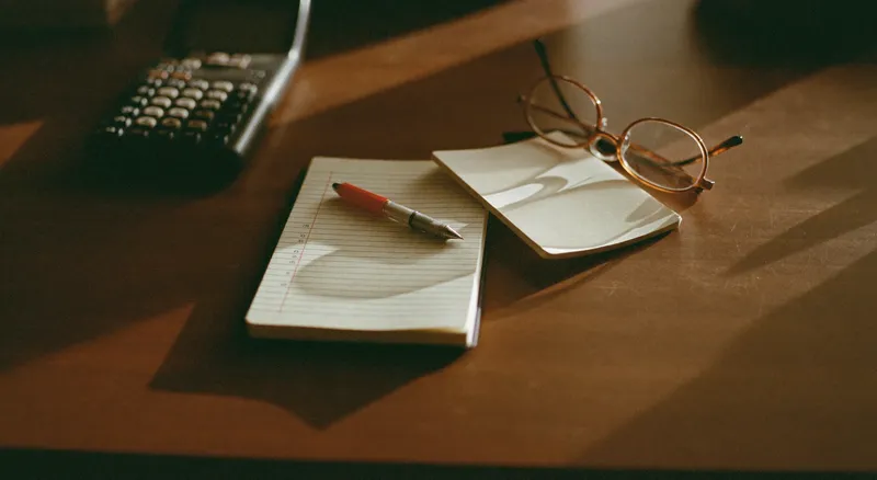 A worn desk calculator, a blank ruled pad, a fountain pen and reading glasses on a timber desk in low afternoon light