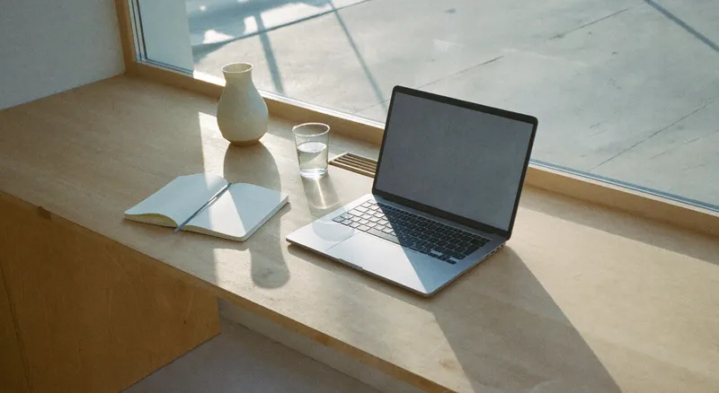 A quiet studio desk beside a large window, laptop open on a page layout, morning light across the timber surface