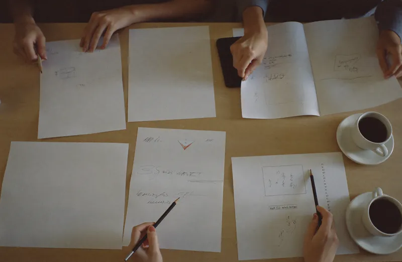 Printed page layouts spread across a plywood table with pencil annotations and two coffee cups