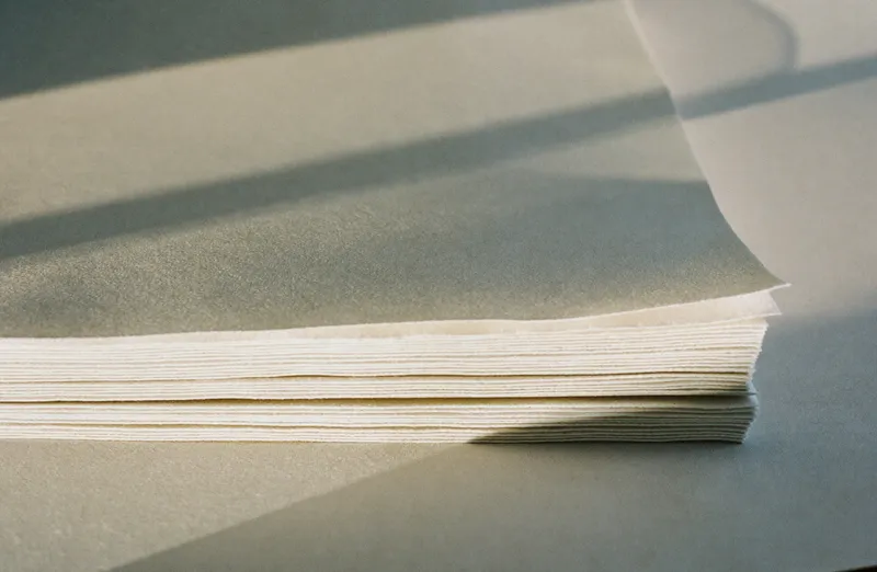 Layered edges of thick off white cotton paper with raking afternoon sunlight casting long soft shadows
